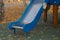 Wooden slide with metal coating, painted blue, standing in playground in park with birch trees and fallen leaves on autumn day