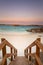 Wooden staircase leading down to a beach at dusk with orange granite rocks in the water and soft color in the sky caused by bush