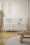 Wooden stool with shoes on round rug in bright baby`s bedroom interior with white cradle