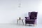 A wooden table and a fancy, dark purple, wing armchair in an empty living room interior with white walls and place for furniture.