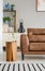 Wooden table next to brown couch in living room interior with plants and striped rug. Real photo