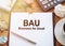 On a wooden table there is an office sheet of paper with the text BAU - Business as Usual. Business workspace with calculator,