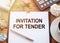 On a wooden table there is an office sheet of paper with the text INVITATION FOR TENDER. Business workspace with calculator,