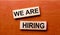 On a wooden table are two wooden blocks with the text question WE ARE HIRING