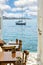 Wooden tables and chairs in a street with a view of a sailboat sailing in the south Aegean sea