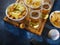 On a wooden tray, two glasses of light beer, onion rings, chips, snacks. There is a remote control and a baseball on the table.