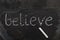 The word BELIEVE written with chalk on black stone