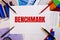 The word BENCHMARK is written on a white background near colored graphs, pens and pencils. Business concept