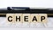 A word Cheap written in a wooden cube with a pen and wallet