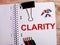 The word CLARITY is written in a notebook near multi-colored pencils and buttons on a wooden background