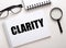 The word CLARITY is written in a white notebook on a white background near a black magnifier and black-framed glasses. Business