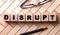 The word DISRUPT is written on wooden cubes on a wooden background next to a pen and glasses