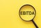 The word EBITDA - Earnings Before Interest, Taxes, Depreciation and Amortization, is written on a magnifying glass on a yellow