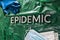 The word epidemic laid by metal letters on crumpled green pastic with face masks and blue protective glove