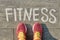 Word fitness written on gray pavement with woman legs in sneakers, view from above