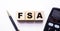 The word FSA Flexible Spending Account is written on wooden cubes between a pen and a calculator on a light background