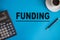 The word funding written on a blue background between a calculator, pen and a cup of coffee
