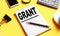 The word GRANT is written in a white notebook on a yellow background near a pen, phone, coffee and other office supplies