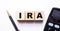 The word IRA Individual Retirement Account is written on wooden cubes between a pen and a calculator on a light background