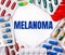 The word MELANOMA is written on a light background surrounded by multi-colored packages with pills. Medical concept