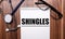 The word SHINGLES is written on white paper on a wooden background near a stethoscope and black-framed glasses. Medical concept