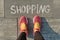 Word shopping written on gray sidewalk with women legs in sneakers, top view