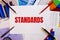 The word STANDARDS is written on a white background near colored graphs, pens and pencils. Business concept