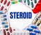 The word STEROID is written on a light background surrounded by multi-colored packages with pills. Medical concept