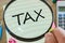 The word `Tax` seen through a magnifying glass.  Next to a notebook, calculator, pen, crumpled balls of paper.