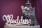 Word `Wedding` made of white wooden letters, statuettes of Eiffel Tower, and cage for birds, stand on table on purple background
