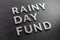 The words rainy day fund laid with silver metal letters on black background, covered with water drops, diagonal perspective,