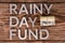Words RAINY DAY FUND laid on wooden surface by metal letters with small roll of us dollar banknotes, flat lay from above
