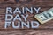 Words RAINY DAY FUND laid on wooden surface by metal letters with small stack of us dollar banknotes