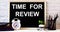 The words TIME FOR REVIEW is written on the chalkboard next to the white alarm clock, glasses, potted plant, and pencils in a