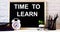 The words TIME TO LEARN is written on the chalkboard next to the white alarm clock, glasses, potted plant, and pencils in a stand