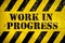 Work in progress warning sign stencil with yellow and black stripes painted over concrete wall cement texture background.