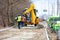 A work team uses a road excavator to unload stacks of paving slabs to the side of the road for subsequent paving