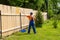 Worker in blue overalls, orange t-shirt, cap and gloves painting a wooden fence