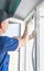 A worker in a blue uniform installs a double-glazed window in a plastic window frame, fixing it with a rubber mallet