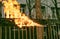 A worker cleans old paint from a metal fence using fire. A man\'s hand holds a gas burner with a lit flame