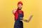 Worker man wearing blue overalls, red T-shirt and cap working on laptop, pointing finger at camera.