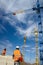 A worker in overalls looks at the high-rise construction cranes on the construction site against a blue sky