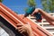 Worker on a roof with electric drill installing red metal tile on wooden house.