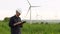 Worker stands near white wind turbines, typing on a tablet. Windmills, green energy concept.