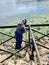 The worker welder works in a protective mask on the shore against the background of water and green aquatic plants