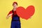 Worker woman pointing at big red heart with finger, looking at camera, expressing positive emotions.