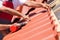 Workers on a roof with electric drill installing red metal tile on wooden house.