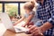 Working Father Using Laptop At Home On Kitchen Counter Whilst Looking After Young Daughter