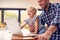 Working Father Using Laptop At Home On Kitchen Counter Whilst Looking After Young Daughter