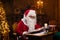 Workplace of Santa Claus. Cheerful Santa is reading the book of wishes while sitting at the table. Fireplace and